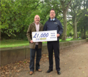 Two men standing outdoors, holding a large cheque for £1,000 from Postcode Dream Trust. One is a renowned taxi sales specialist. Trees and a park can be seen in the background.