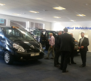 Business professionals converse inside a modern showroom showcasing multiple taxis. The wall features signage for Allied Vehicles and Cab Direct, creating a professional and energetic setting ideal for taxi sales.