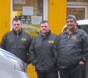 Three men in black jackets stand together outside a bright yellow building next to a parked Renault Trafic taxi.