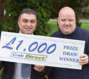 Two men stand outdoors, smiling and holding a large sign that reads £1,000 Cab Direct PRIZE DRAW WINNER. Greenery and trees form the background, highlighting their excitement as proud winners from Cab Direct’s taxi prize draw.