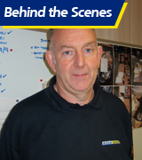 A man stands in front of a whiteboard with notes and photos, demonstrating the taxi quality control process. He wears a dark shirt with a logo, and “Behind the Scenes” appears at the top in white and yellow text.