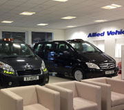 Two black taxis and sleek black cabs are displayed in a showroom with beige armchairs in the foreground and an Allied Vehicles sign on the back wall.