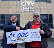 Two men shake hands outside a City Taxis building, with one holding a sign that says £1,000 Prize Draw Winner Cab Direct, celebrating his recent prize win—ideal for promoting taxi sales and success stories.