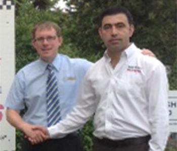 Two men stand outdoors, shaking hands during a taxi prize draw announcement. One wears glasses, a blue shirt, and striped tie; the other is in a white shirt. Green foliage and part of a sign appear in the background.