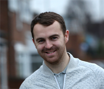A young man with short brown hair and a trimmed beard smiles outdoors in a light grey zip-up jacket, standing against a blurred urban background, appearing happy—possibly after winning a taxi prize draw.