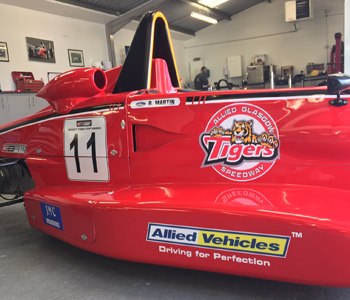 A red Formula Ford racing car featuring number 11 and Allied Glasgow Tigers Speedway along with Allied Vehicles sponsor logos, displayed indoors with people and promotional images in the background.