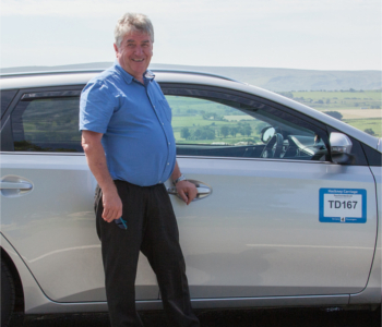 A man in a blue shirt stands smiling next to a silver Auris Touring taxi parked outdoors, with a green landscape in the background and a car door sign reading TD167.