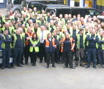 A group of workers in high-visibility vests pose for a group photo in an industrial area next to a fleet of bright yellow taxis.