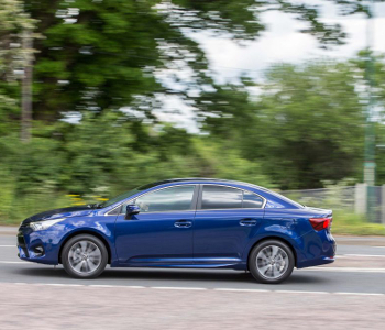A blue Toyota Avensis saloon taxi drives swiftly along a road, with blurred green trees in the background highlighting its smooth performance and reliability.