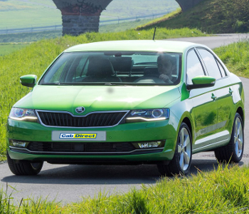 Green saloon car available for taxi service driving on a rural road, featuring green grass and a stone bridge in the background. Driver is visible through the windscreen.
