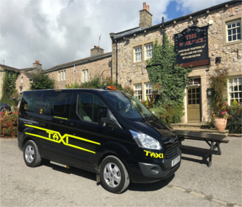 A Ford ProCab™ black taxi with yellow markings is parked outside a stone building with a sign reading The Woolpack, surrounded by greenery and a wooden picnic table.