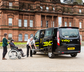 A taxi driver assists a wheelchair user boarding a black Top Track 250 taxi via ramp, whilst two people stand nearby in front of a historic building.