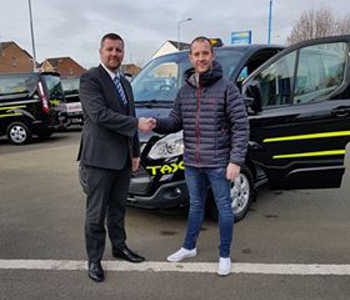 Two men shake hands in a car park next to a black taxi, marking ProCab’s South Ayrshire launch. One man is dressed in a suit, the other in a casual jacket and jeans. Other taxis and houses appear in the background.