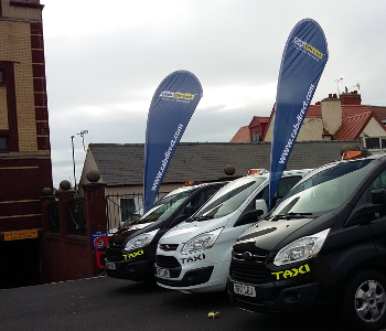 Three taxis, two white and one black, are parked side by side with blue COAST taxi advertising flags behind them, showcasing their affiliation with the National Taxi Association. Buildings with red roofs are visible in the background.