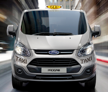 A white Ford taxi with a yellow taxi sign on top drives through a city street, highlighting its motion and reliability as part of the Taxi Roadshow.