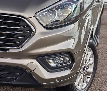 Close-up of the front right side of a metallic bronze Ford Tourneo Custom taxi, highlighting the headlamp, grille, fog lamp, and part of the alloy wheel on a tarmacked road.