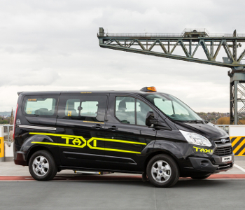 Black taxi with yellow stripes and TAXI sign, parked outdoors near industrial equipment under a cloudy sky—reliable choice for professional taxi services.