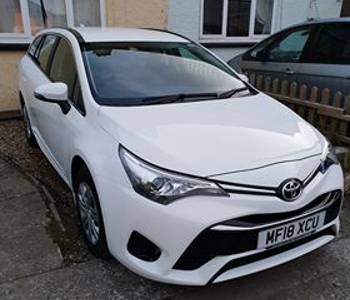 A white Toyota Avensis Tourer estate taxi with registration plate MF18 XCU is parked beside a house, next to a small fence and another car.