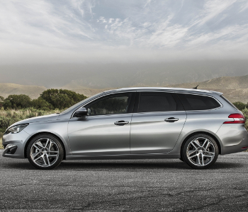 A silver Peugeot 308 estate car taxi is parked on a paved road with scenic mountains and greenery in the background under a cloudy sky.