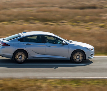 A New Generation Vauxhall silver saloon taxi drives swiftly on a tarmacked road through a dry, grassy landscape, with motion blur in the background.