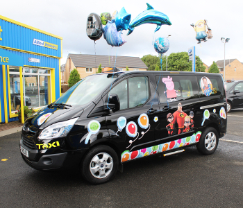 A black taxi featuring colourful cartoon character stickers and balloons, including a dolphin, parked outside a building with yellow and blue signage—an eye-catching and unique taxi ideal for attracting attention or special events.