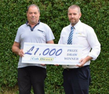 Two men stand outside holding a large sign announcing the winner of the £1,000 Cab Direct prize draw, facing the camera with a leafy green hedge behind them.