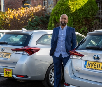 A man in a blue suit stands between two silver Toyota Auris Icon-Tech cars outside, set against greenery and a brick building—ideal vehicles for any taxi fleet.