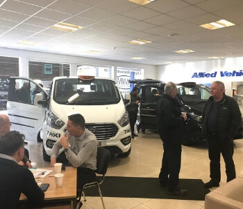 Visitors engage in conversations inside a car showroom featuring several taxis on display. Two men stand and chat near a white taxi, while others talk at a nearby table. The Allied Vehicles sign is visible, capturing the bustling environment of a taxi roadshow event.
