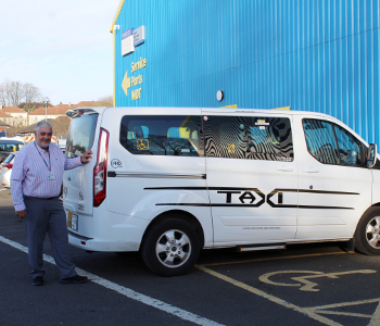 A man stands beside a white ProCab™ wheelchair-accessible taxi parked in a designated accessible parking bay near a blue building with yellow signs.