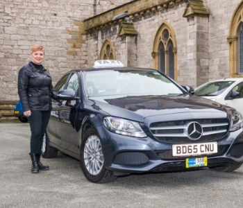 A woman in a black jacket stands next to a dark grey Mercedes taxi, showcased among several quality used taxis parked by a stone building with arched windows, ideal for professional taxi services.