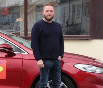 A red Ford Mondeo taxi parked by a building with large windows, with a bearded man in a dark jumper and jeans standing beside it.