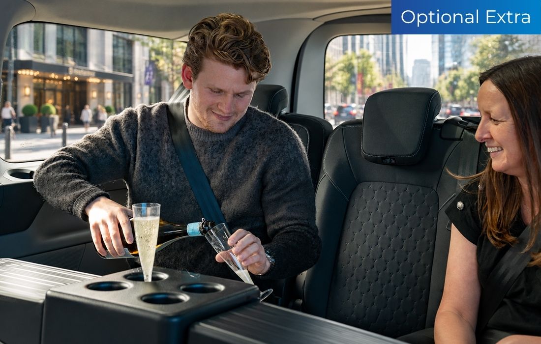 A man pours champagne into a glass while seated in the back of a Ford Tourneo Custom Taxi beside a smiling woman. An “Optional Extra” sign is displayed in the top right corner.