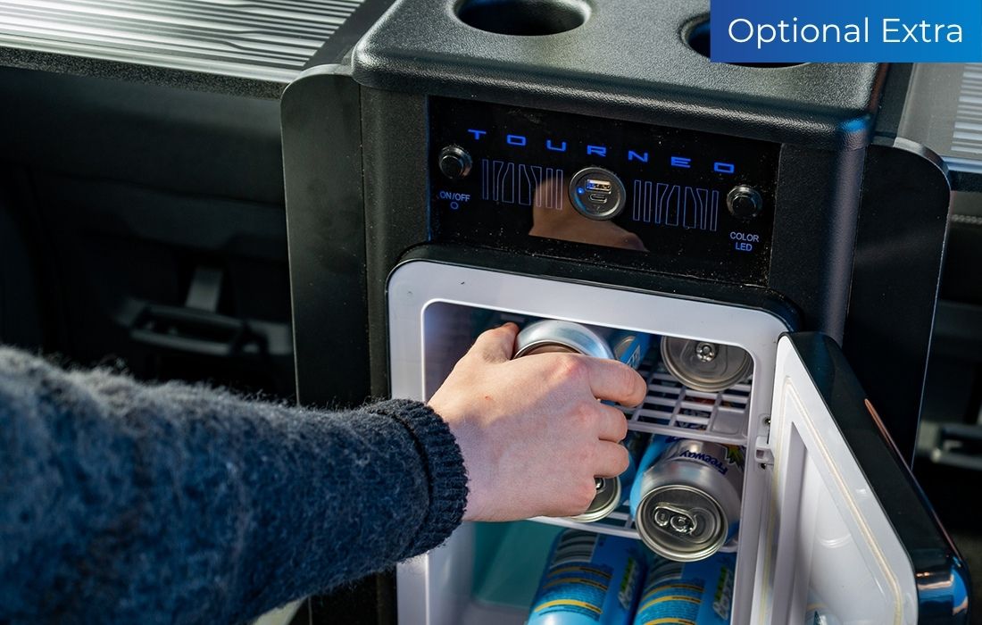 A person’s hand reaches into a small fridge inside a Ford Tourneo Custom Taxi to grab a can, with other cans visible. The upper right corner displays an “Optional Extra” label, highlighting premium taxi features.