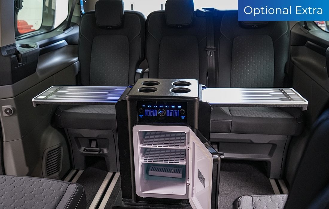 Ford Tourneo Custom Taxi interior featuring three rear seats, an open small fridge, cup holders, and fold-out trays for enhanced passenger comfort. A blue label in the top right corner reads “Optional Extra”.