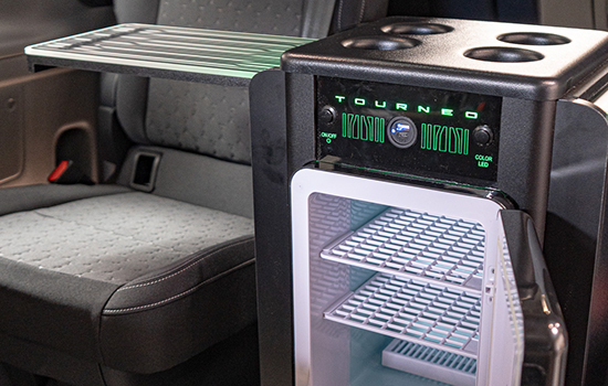 A compact mini fridge with a transparent door and shelves is installed inside a Ford Tourneo Custom taxi, positioned next to a folded tray and car seat, and labelled “Tourneo” on the front panel, enhancing passenger comfort and convenience.