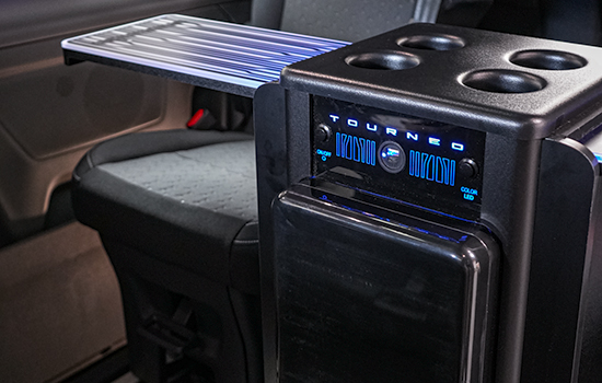 Premium interior of the Ford Tourneo Custom Taxi showcasing a fold-out tray, integrated cup holders, and a control panel with blue LED lights labelled Tourneo. Adjacent seat features durable grey fabric upholstery.