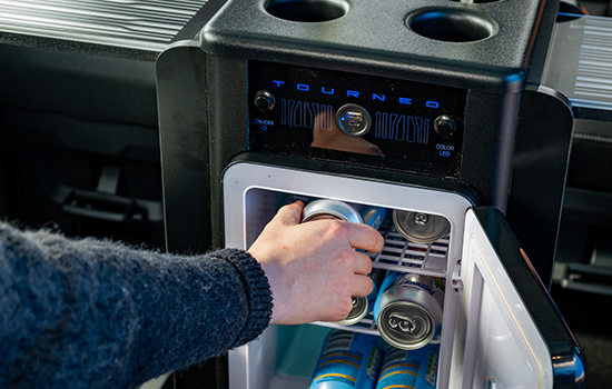 A person’s hand reaches into a small fridge stocked with tins inside a taxi. The fridge is built into a console labelled Tourneo with cup holders on top, showcasing the premium amenities available in the Ford Tourneo Custom Taxi.
