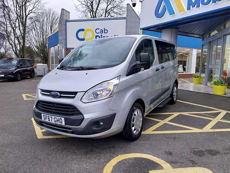 A silver Ford taxi parked in a disabled parking bay outside a building with Cab Direct and Motability signs. Trees and another car are visible in the background.