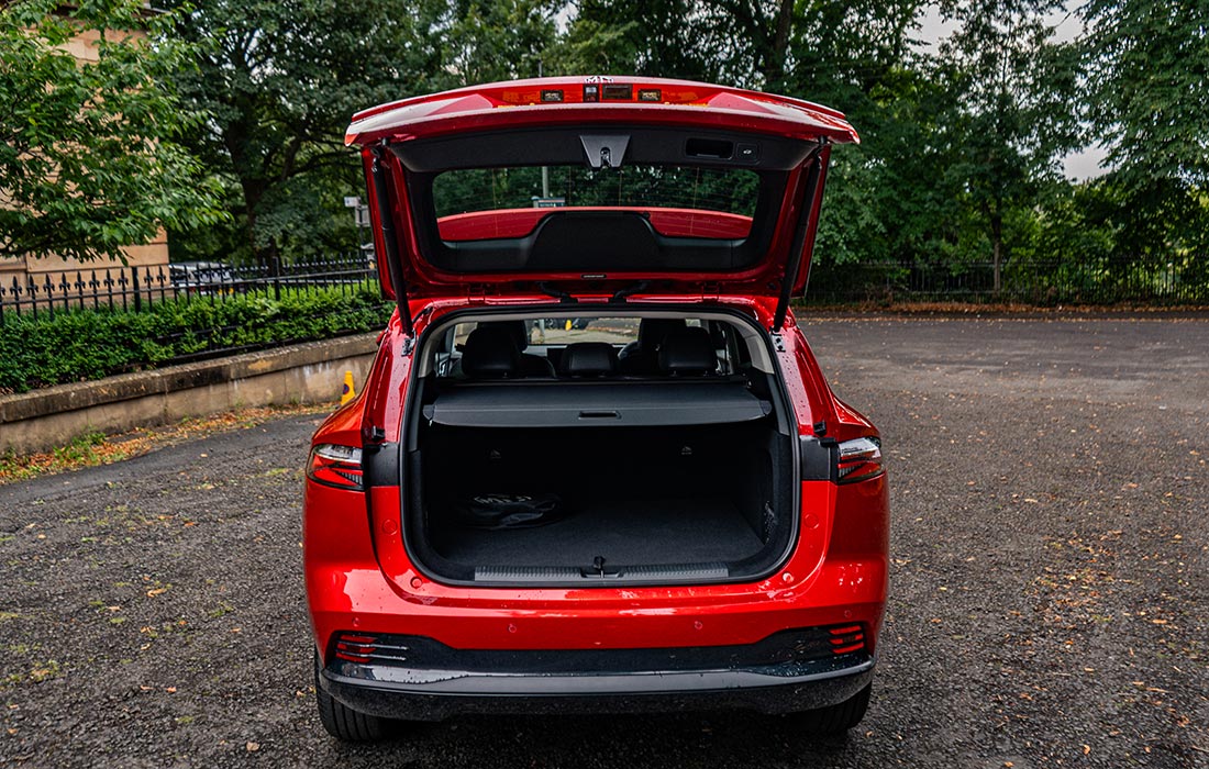 A red SUV taxi with its rear hatch open, displaying a spacious empty boot. The vehicle is parked on a gravel car park with trees and a metal fence in the background.