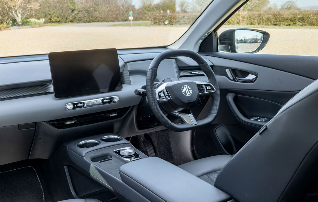 Sleek MG S5 EV interior featuring a digital display, MG logo steering wheel, convenient cup holders, and easy-to-use control knobs. The black and grey colour scheme gives a contemporary look ideal for commercial taxi use. Outside landscape visible through the windows.