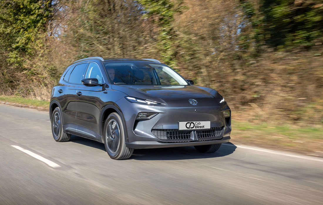 A sleek, dark grey MG S5 EV electric SUV taxi drives on a sunny road lined with trees and bushes. Modern styling and a distinctive front with prominent headlamps make it an ideal taxi for comfort and efficiency.
