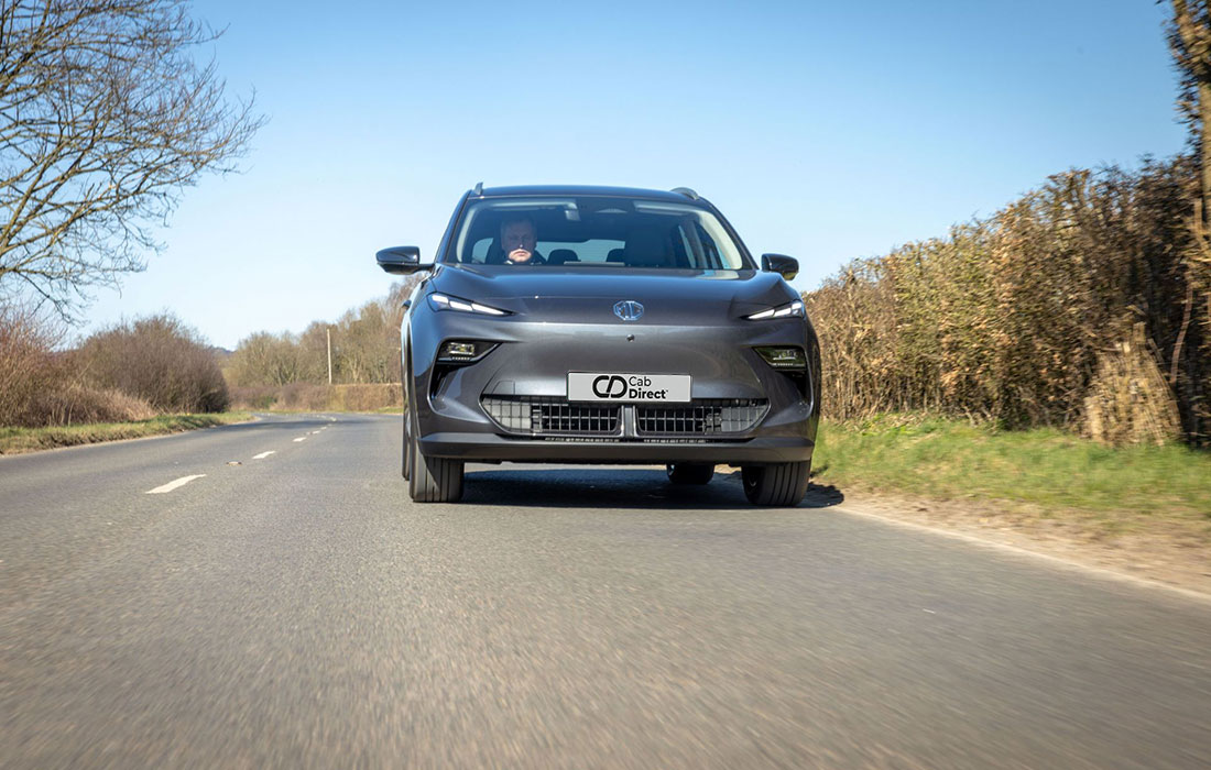 A grey MG S5 EV SUV taxi drives on a rural road lined with leafless trees and hedges beneath a blue sky, featuring a Car Direct plate on the front grille.