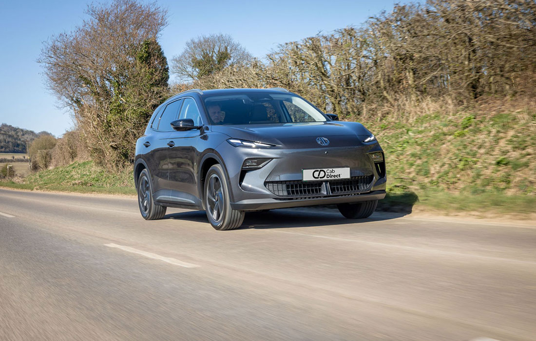 A sleek grey MG S5 EV electric SUV drives on a country road with lush greenery and a clear sky, showcasing its style and suitability for taxi fleets.