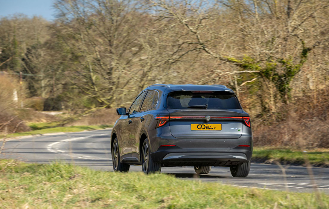 A grey MG S5 EV SUV taxi drives on a winding rural road lined with grass and leafless trees, beneath a clear sky.