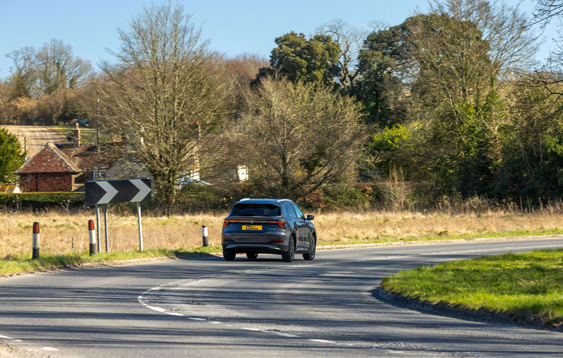A black MG S5 EV taxi drives away on a curving country road, with grass, trees, and houses in the background under a clear sky.