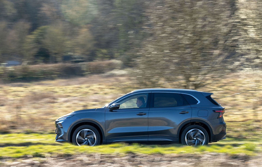 A silver MG S5 EV SUV taxi drives through a grassy field, side view captured with motion blur in the background and sunlight highlighting the landscape.