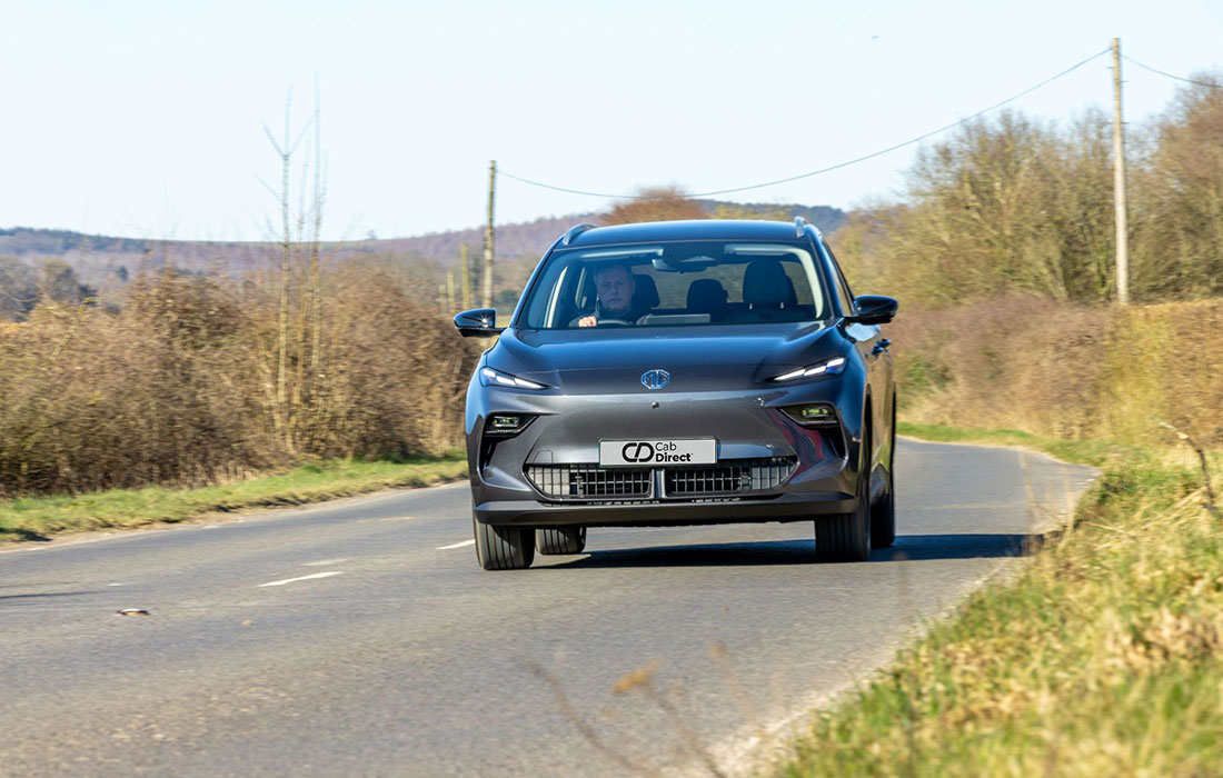 A grey MG S5 EV electric SUV taxi drives along a rural two-lane road lined with bushes and grassy areas, with leafless trees and hills in the distance under a clear sky.
