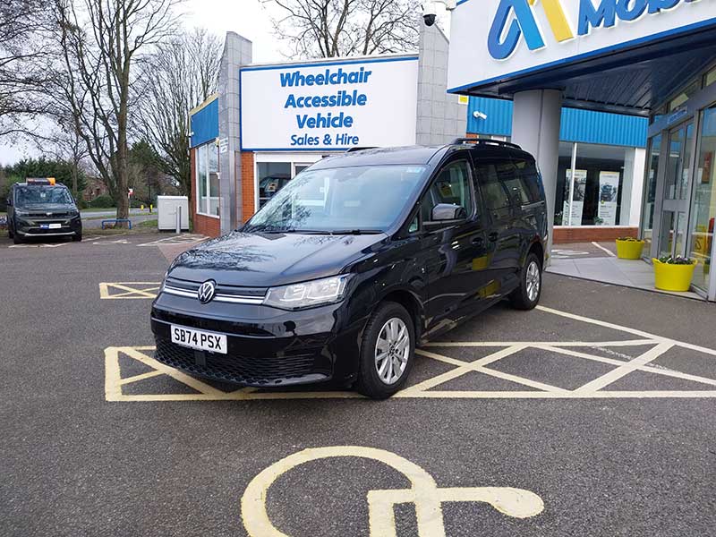 A black Volkswagen wheelchair accessible taxi is parked in a disabled parking bay outside a building with a sign reading Wheelchair Accessible Vehicle Sales & Hire.