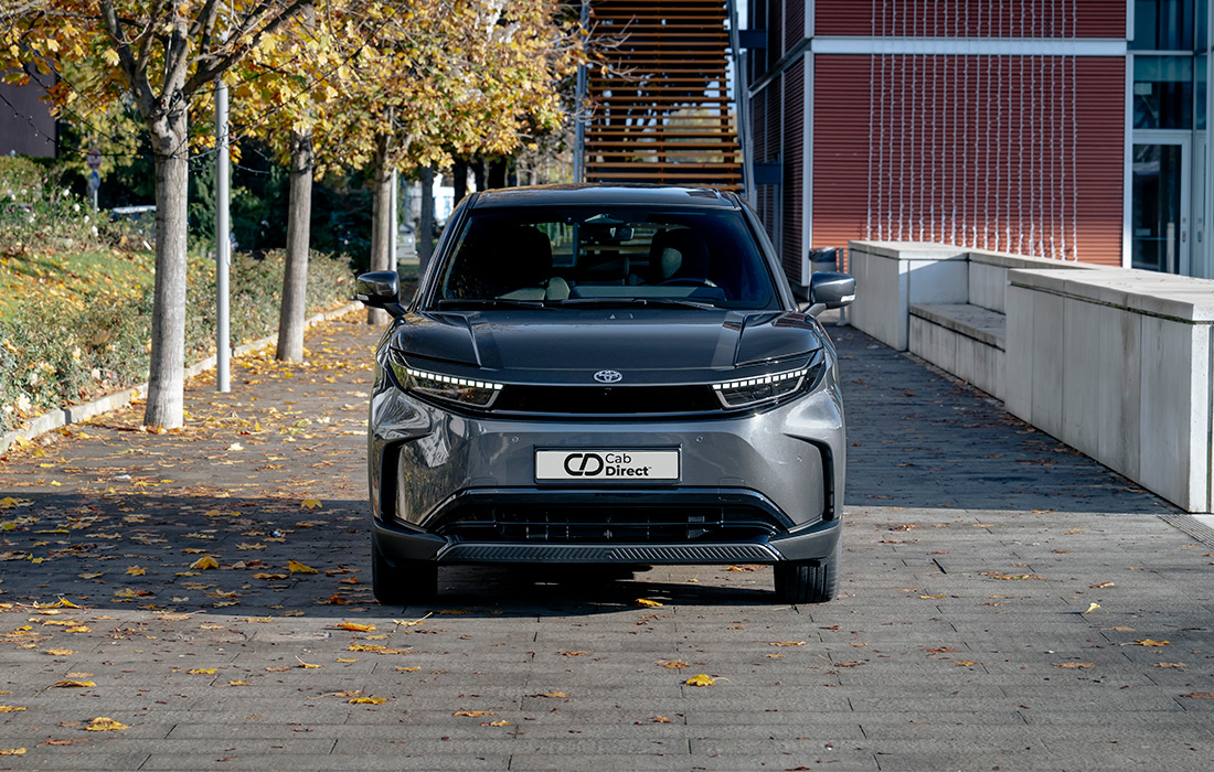 A grey Toyota SUV taxi is parked on a leaf-strewn pavement in front of modern buildings and autumn trees, front view.