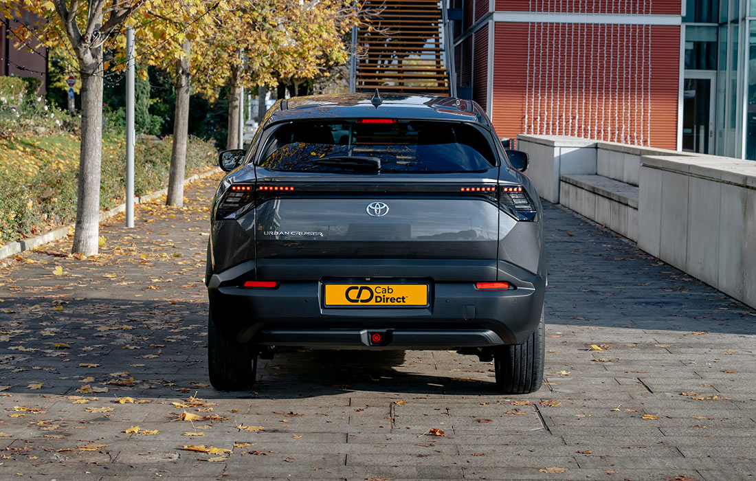A grey Toyota taxi is parked on a paved path lined with autumn trees, rear view visible with a Cab Direct number plate. Modern buildings and stairs are in the background.